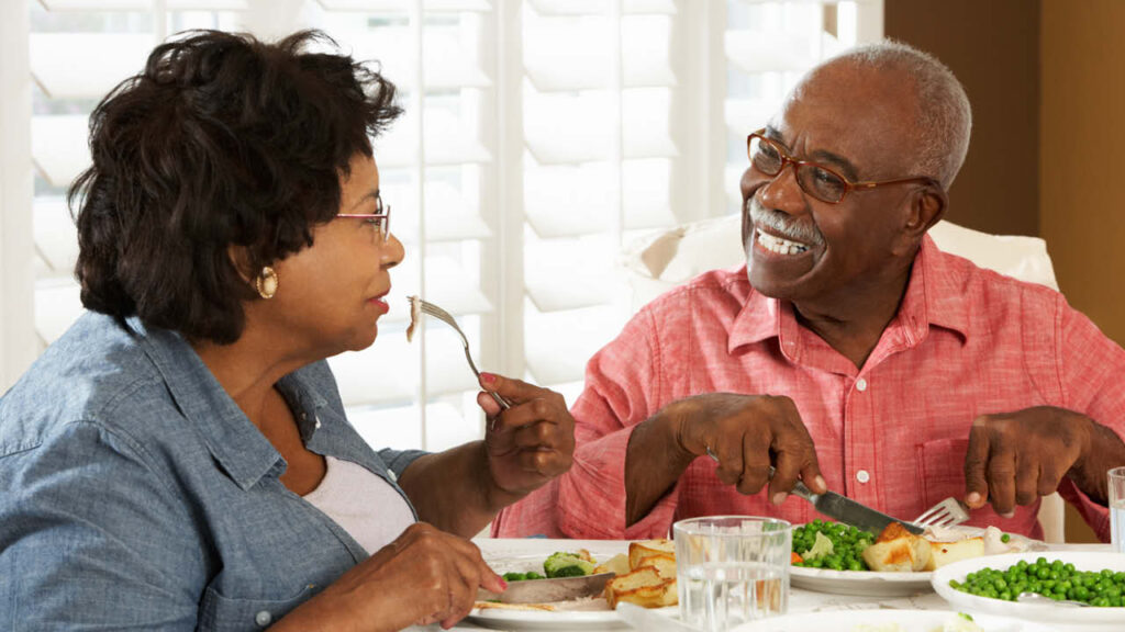 An older couple sits at a dining table, smiling and enjoying a meal together. The man wears glasses and a pink shirt, while the woman wears a blue shirt. Plates of food and a glass of water are in front of them.