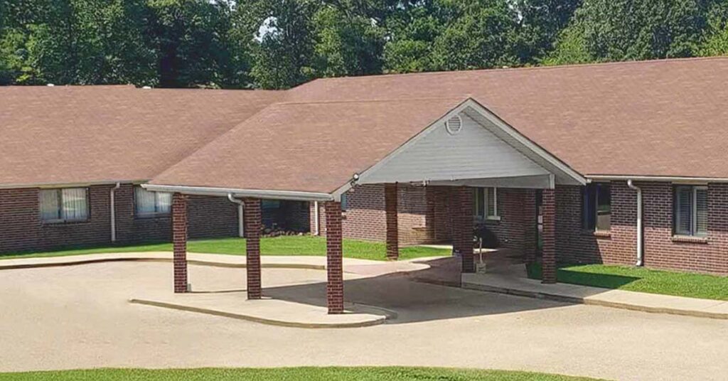 A single-story brick building with a covered driveway entrance supported by brick columns, surrounded by green grass and trees in the background.