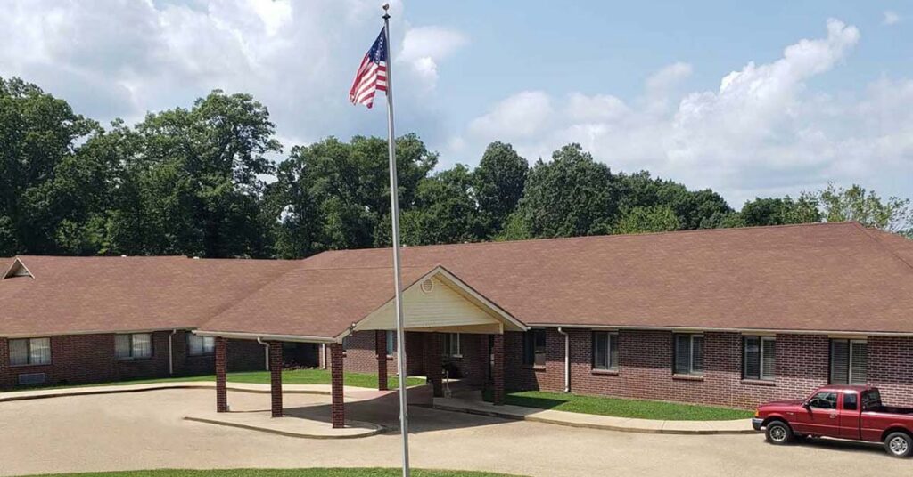 A brick building with a brown roof and covered entrance sits behind a circular driveway. An American flag is raised on a flagpole in front, and a red pickup truck is parked nearby. Green trees and blue sky are in the background.