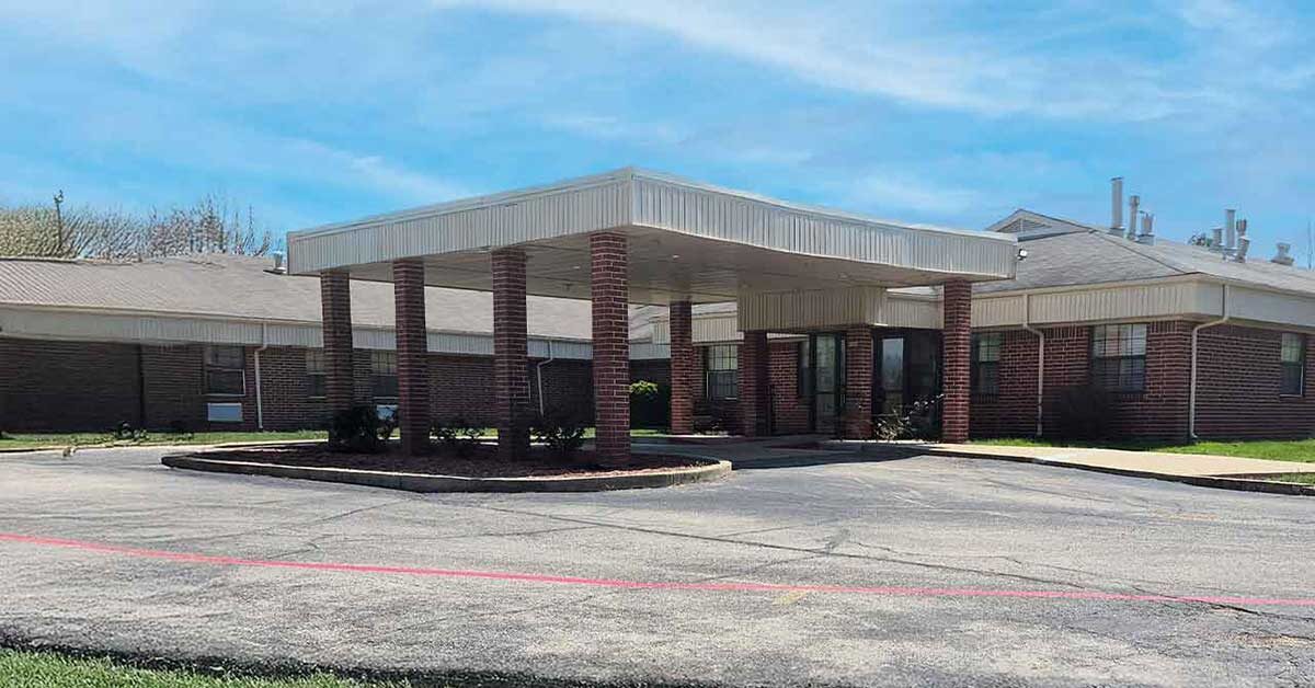 Single-story brick building with a covered entrance supported by brick columns, a circular driveway, and a cloudy blue sky in the background. The area around the building is paved and landscaped with some greenery.