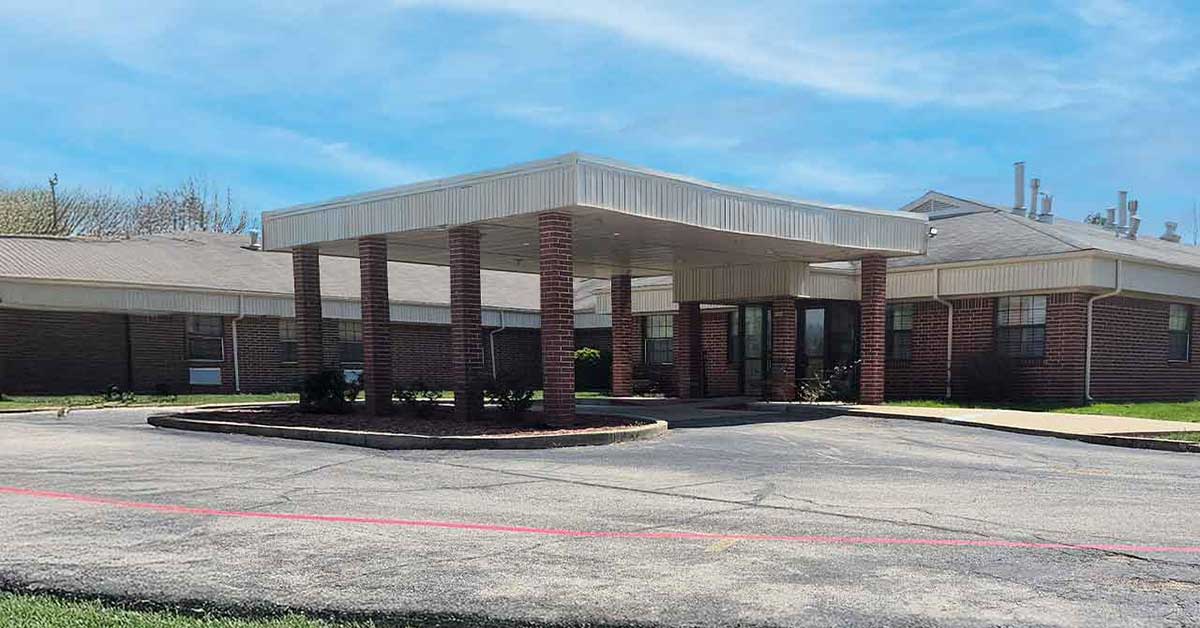 Single-story brick building with a covered entrance supported by brick columns, a circular driveway, and a cloudy blue sky in the background. The area around the building is paved and landscaped with some greenery.