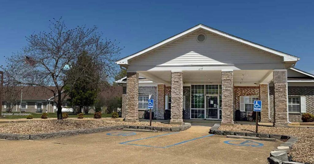 Single-story building with stone columns and a covered entrance, featuring two accessible parking spaces in front. There are leafless trees, a U.S. flag, and clear blue sky in the background.