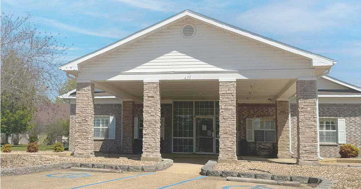Single-story building with stone columns, white trim, and large front entrance. There are windows on either side, a gravel landscape, and wheelchair-accessible parking spaces in front. The sky is partly cloudy.