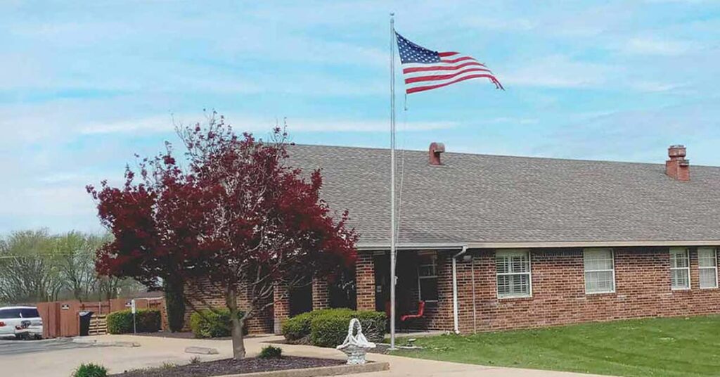 A brick house with a grey roof, a front porch, and an American flag flying on a pole. A small tree with red leaves and a white statue are in the yard, with a car parked on the left side.