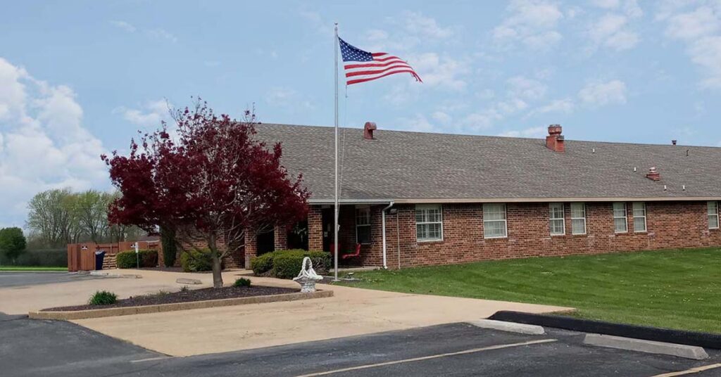 A single-story brick building with an American flag on a tall flagpole in front, surrounded by green grass, a red-leaved tree, and a paved driveway on a partly cloudy day.