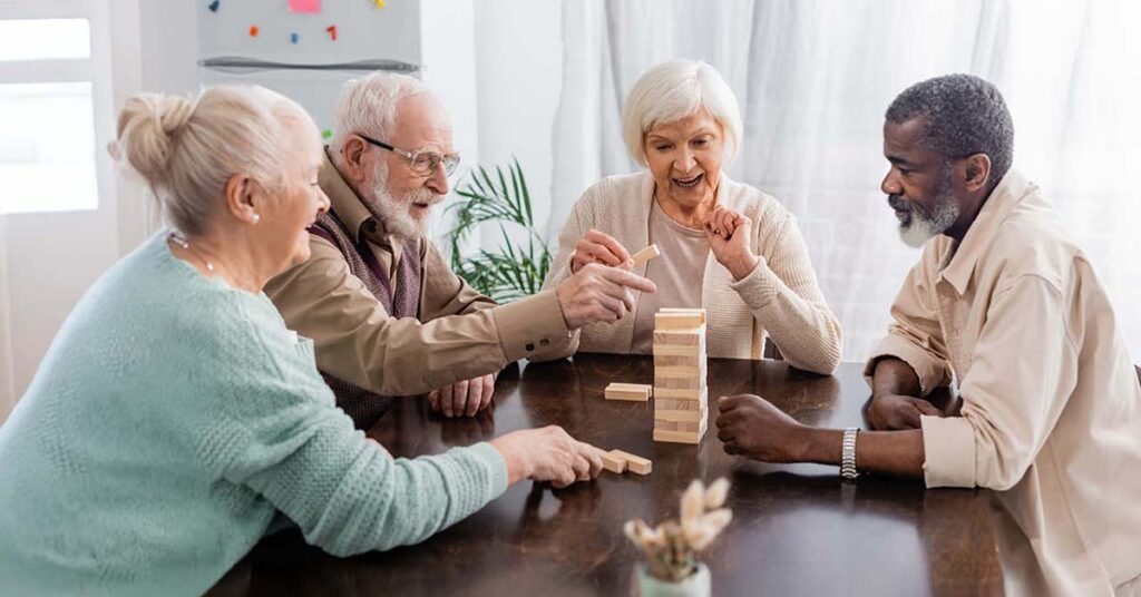 Four older adults sit around a table, smiling and playing a game of Jenga together in a bright, cozy room.