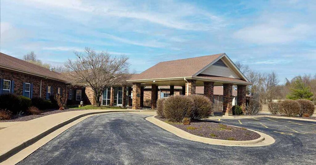 A single-story brick building with a covered entrance and driveway, surrounded by trimmed bushes and bare trees, under a partly cloudy sky.