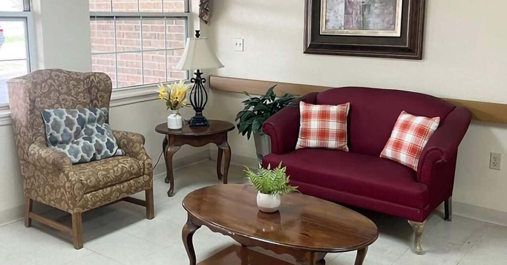 Cozy sitting area with a patterned armchair, maroon loveseat with plaid pillows, wooden coffee and side tables, table lamp, potted plants, and large window letting in natural light.