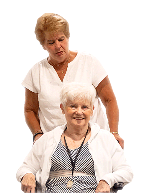 Two older women, one with short white hair sitting in a wheelchair and smiling, the other standing behind her with a gentle expression—both wearing light-colored tops—portray the warmth and companionship found in senior living, set against a white background.