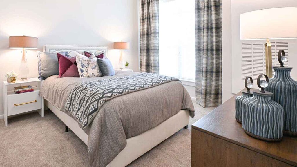 A bright, modern bedroom with a neatly made bed featuring patterned pillows and a gray bedspread, flanked by two white nightstands with lamps. Decorative vases sit on a wooden dresser in the foreground.