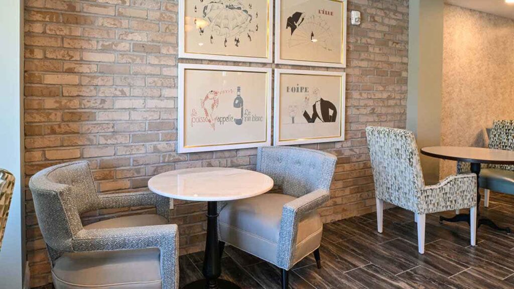 A cozy café corner with a round marble table, two cushioned chairs, and framed artwork on a brick wall, next to another seating area with patterned chairs on a tiled floor.