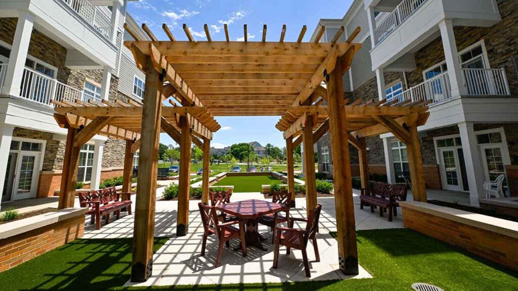 A wooden pergola with seating underneath stands in a sunny courtyard between two modern apartment buildings, surrounded by green lawns and landscaping.