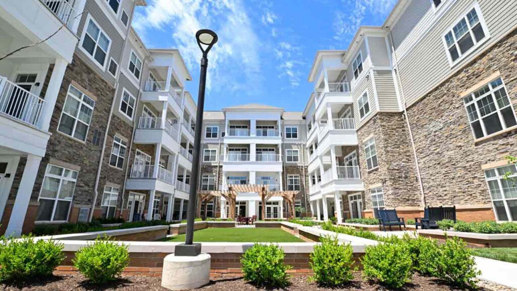 Modern apartment complex courtyard with green landscaping, benches, and a central lamp post, surrounded by multi-story buildings with balconies and stone accents under a bright blue sky.