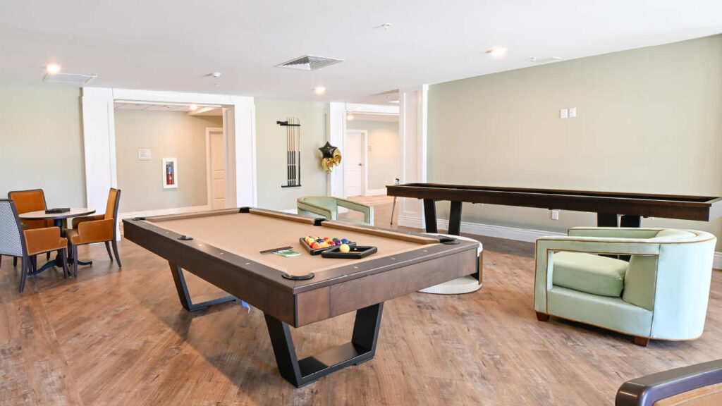 A spacious game room with a pool table, a shuffleboard table, a card table with four chairs, and a green armchair on a wood floor with light-colored walls.