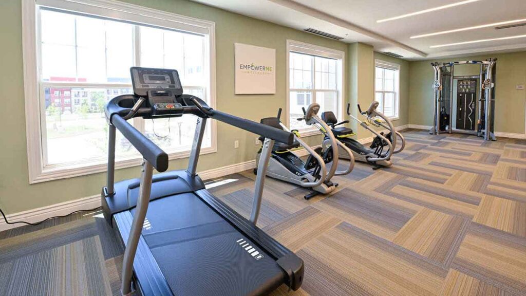 A bright fitness room with large windows, featuring a treadmill, two stationary bikes, and a multi-function cable machine on patterned carpet flooring.