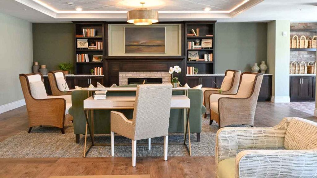A cozy lounge with armchairs and a table arranged around a fireplace, bookshelves on either side, and decorative baskets along the wall; neutral tones and soft lighting create a welcoming atmosphere.