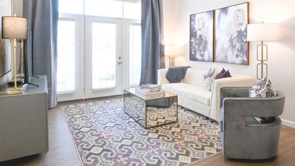 Bright living room with large windows, a cream sofa, gray armchair, glass coffee table, patterned rug, and two abstract floral paintings on the wall. Floor lamps and decorative pillows complete the modern decor.
