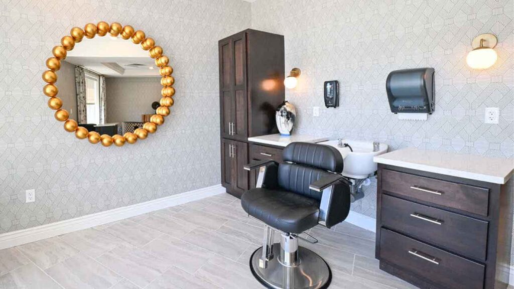 Modern salon room with a black styling chair, dark wood cabinets, a sink, gold circular mirror, wall sconce, paper towel dispenser, and patterned wallpaper. Light wood floor tiles and bright natural lighting.