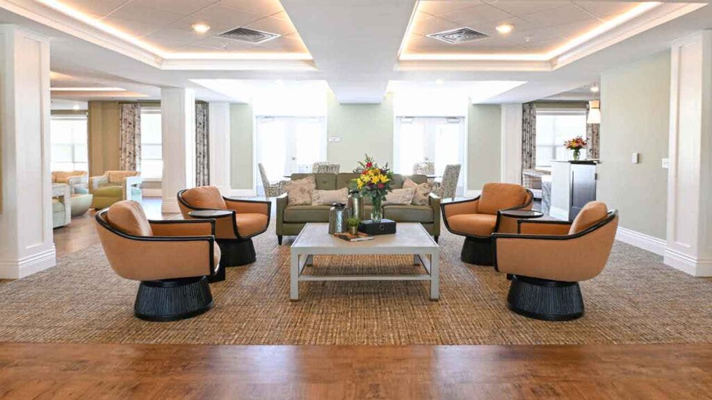 A spacious, modern lounge with four tan swivel chairs around a coffee table, a beige sofa in the background, neutral walls, large windows, and soft ceiling lights. A vase of flowers sits on the table.