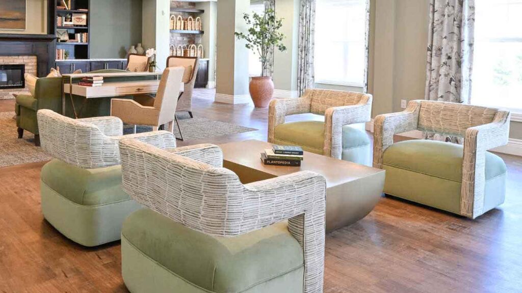 Modern lounge area with four light green and white woven armchairs around a sleek coffee table, set on wooden flooring. A desk, shelves, and decorative plants are visible in the background. Natural light fills the room.