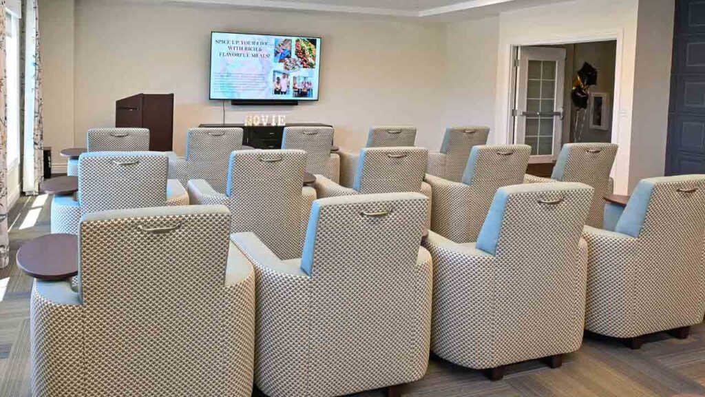 A small conference room with rows of cushioned chairs facing a screen displaying a presentation. There are side trays attached to each chair and a podium in the front left corner. The room has neutral tones and ample lighting.