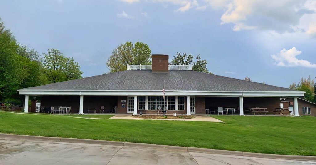 A single-story brick building with a wide, dark roof and a central chimney, set on a grassy lawn with scattered outdoor seating and an American flag in front, surrounded by green trees under a partly cloudy sky.
