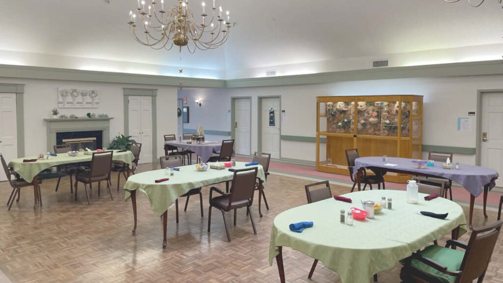 A dining room with several spaced-apart tables, each set with chairs, napkins, and small centerpieces. A chandelier hangs from the ceiling, and a large display cabinet is against one wall. The room is well-lit and tidy.