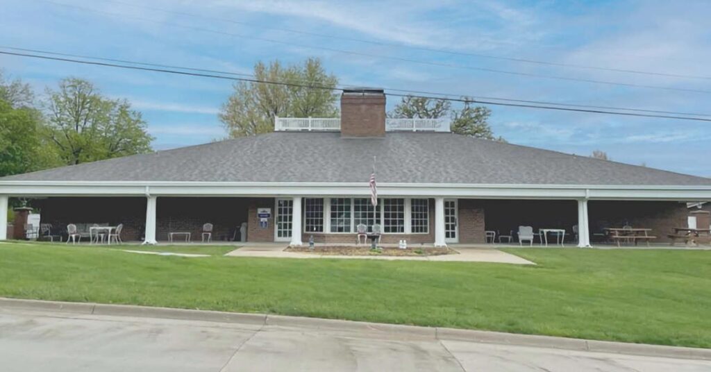 A single-story brick building with a wide porch, several chairs and tables, an American flag, and a small garden in front, set against a backdrop of trees and a blue sky with scattered clouds.