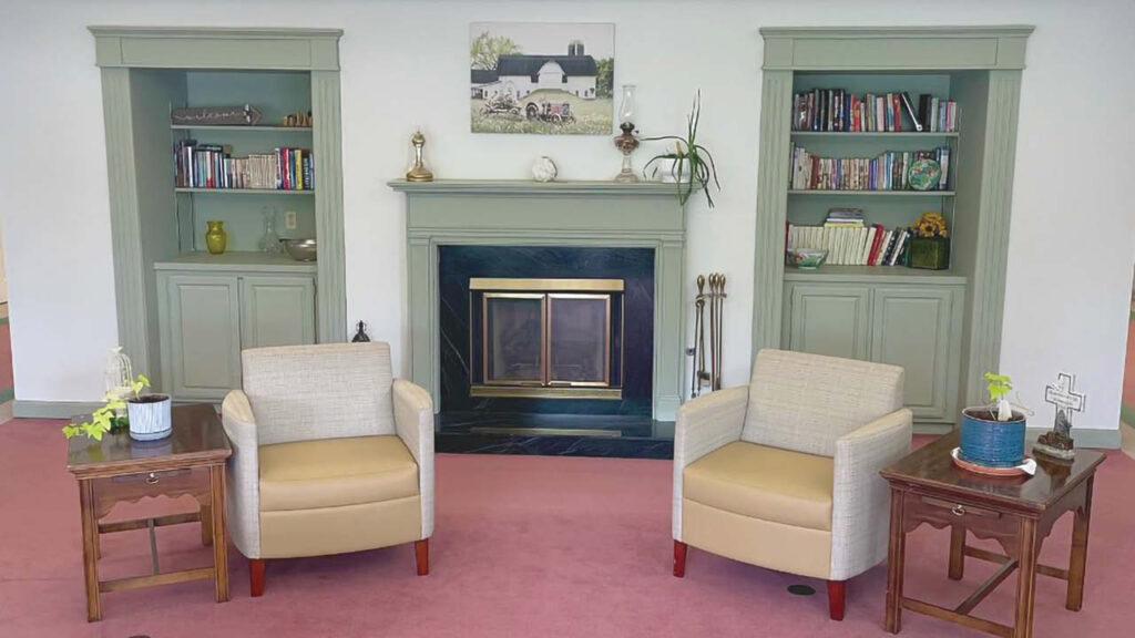 Two beige armchairs sit in front of a fireplace with a black and green mantel, flanked by green built-in bookshelves. Small tables with potted plants are beside each chair, and a countryside painting hangs above the fireplace.
