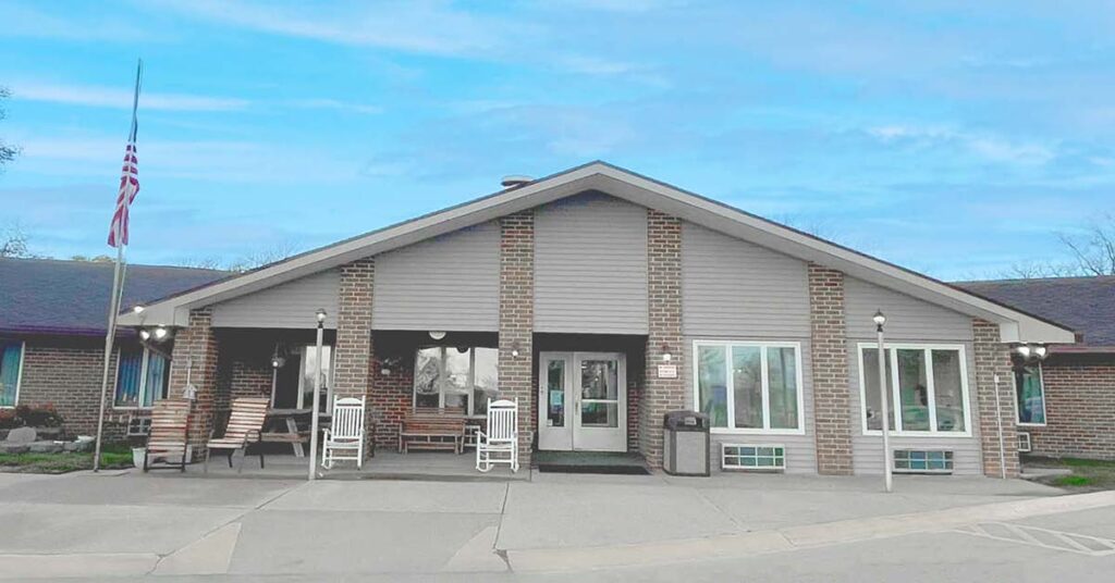A single-story brick building with a covered entrance, several windows, rocking chairs on the porch, and a U.S. flag at half-mast, under a blue sky.