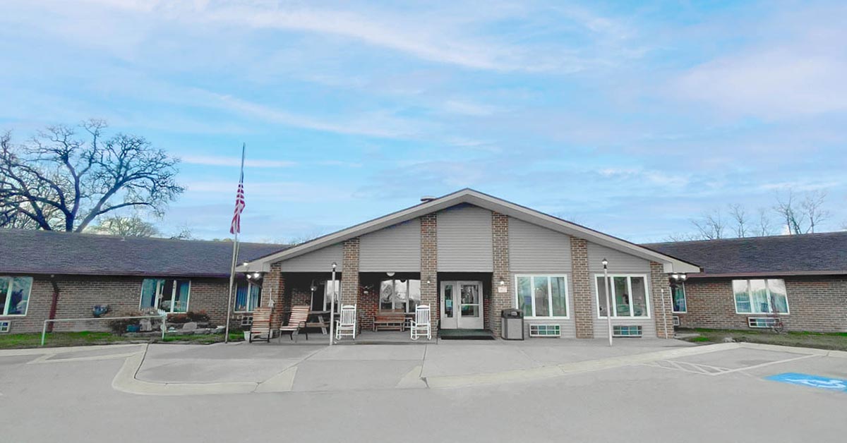 Single-story brick building with a covered entrance and rocking chairs on the porch. An American flag is at half-mast outside. The sky is mostly clear with some clouds. Accessible parking spaces are visible in front.