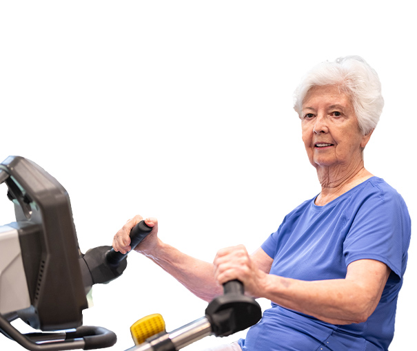 An older woman with short white hair, wearing a blue shirt, is using a stationary exercise machine and smiling at the camera, showcasing active living in an Assisted Living environment against a white background.