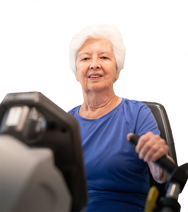 An elderly woman with white hair and a blue shirt is smiling while using exercise equipment, holding the handles and looking forward. The background is white.