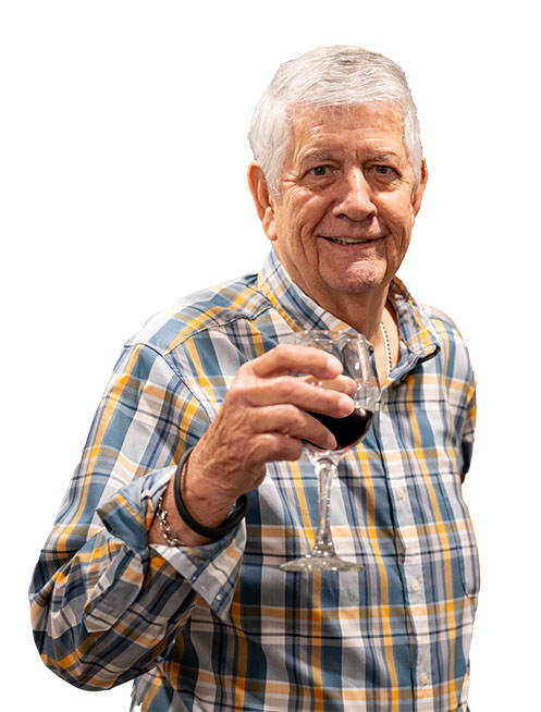 An older man with gray hair, dressed in a plaid shirt, smiles while holding a glass of red wine and raising it in a toast—celebrating the joys of Independent Living against a clean white background.