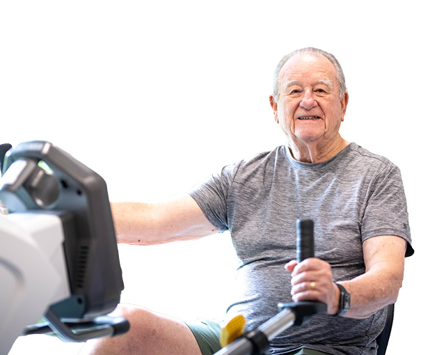 An older man in a gray t-shirt and shorts smiles while using a rowing machine, exercising indoors. The brightly lit, mostly white background highlights his commitment to fitness and Independent Living.