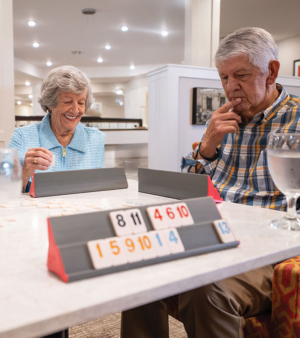 Two elderly people sit at a table playing a tile-based game, smiling and concentrating. Game racks with numbered tiles are in front of each person. Water glasses are on the table in a warmly lit room.