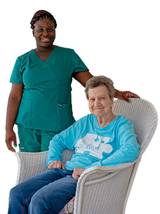A smiling healthcare worker in green scrubs stands beside an elderly woman seated in a white wicker chair, both sharing a joyful moment—a glimpse into the rewarding world of Senior Living Careers.