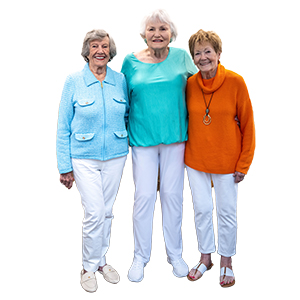 Three older women stand together smiling, embodying the joy of senior living. One wears a light blue jacket, one a teal top, and one an orange sweater; all wear white pants and light shoes. The background is plain white.