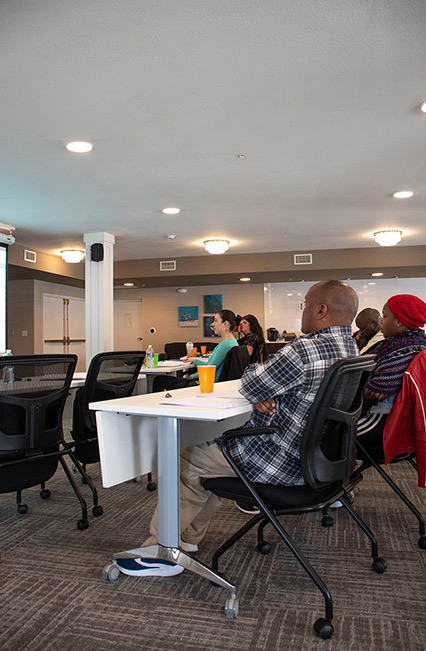 People sit at tables in a conference room, attentively watching a presentation projected onto a screen at the front, with drinks and notebooks on the tables. The room has modern decor and bright lighting.