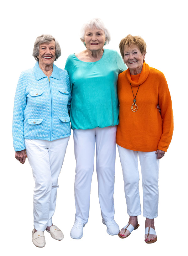 Three smiling older women stand together. One wears a blue jacket, another a turquoise top, and the third an orange sweater; all are wearing white pants. The background is plain white.
