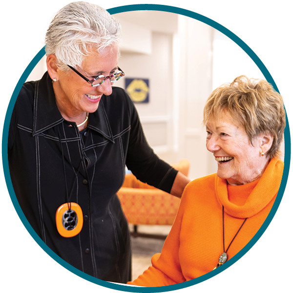 Two older women smiling and talking; one is standing and wearing glasses and a black top with an orange necklace, the other is seated, wearing an orange sweater. They appear happy in a bright indoor setting.