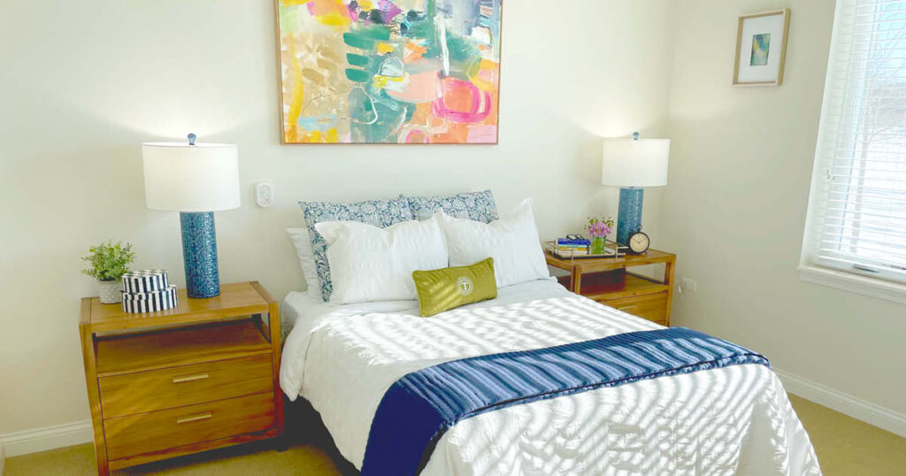 A bright bedroom with a neatly made bed, blue-and-white pillows, wooden nightstands with lamps, and a colorful abstract painting above the headboard. Sunlight filters through window blinds, casting striped shadows on the bed.