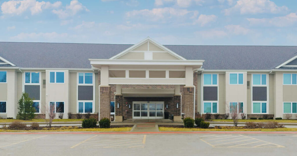 A large, two-story building with a covered entrance, brick and beige siding, surrounded by small shrubs and a mostly empty parking lot under a partly cloudy sky.