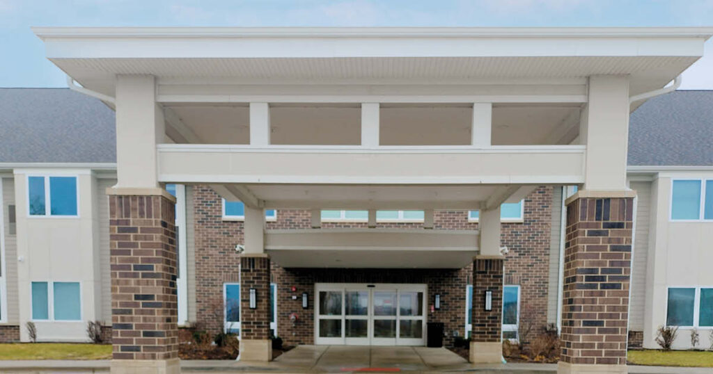 A brick and siding building with a covered entrance supported by columns, large windows, and glass double doors, likely the entrance to a modern commercial or healthcare facility.