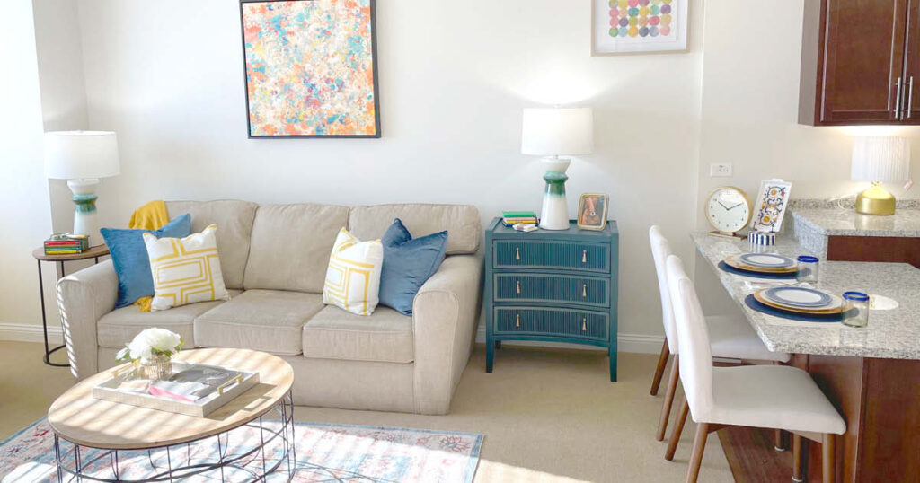 Bright living room with a beige sofa, colorful cushions, coffee table with flowers and books, blue dresser, wall art, two lamps, and a breakfast bar set with plates and chairs. Light and inviting decor.