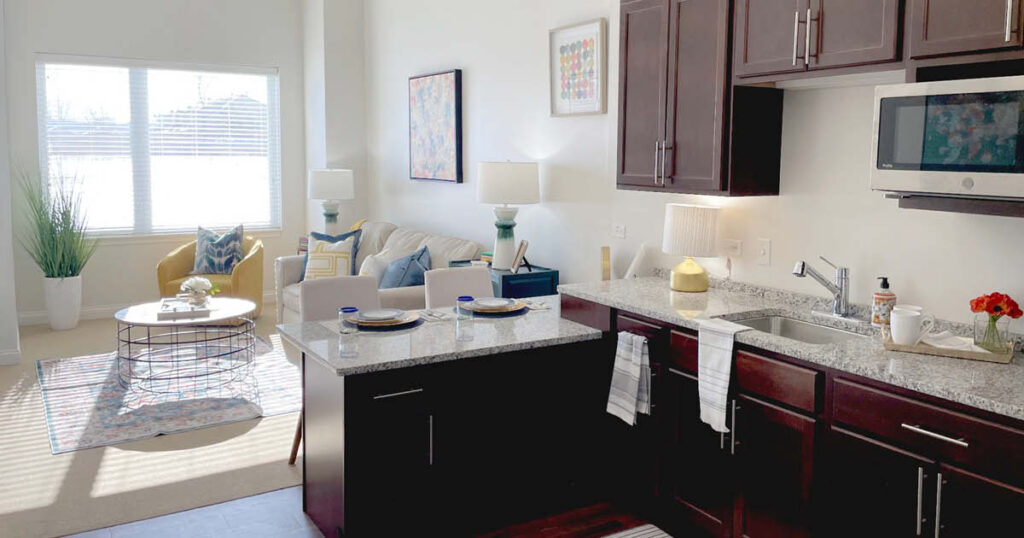 A modern, bright apartment kitchen with dark cabinets, granite countertops, and a breakfast bar set for two, opening to a sunlit living area with a sofa, coffee table, armchair, and large window.