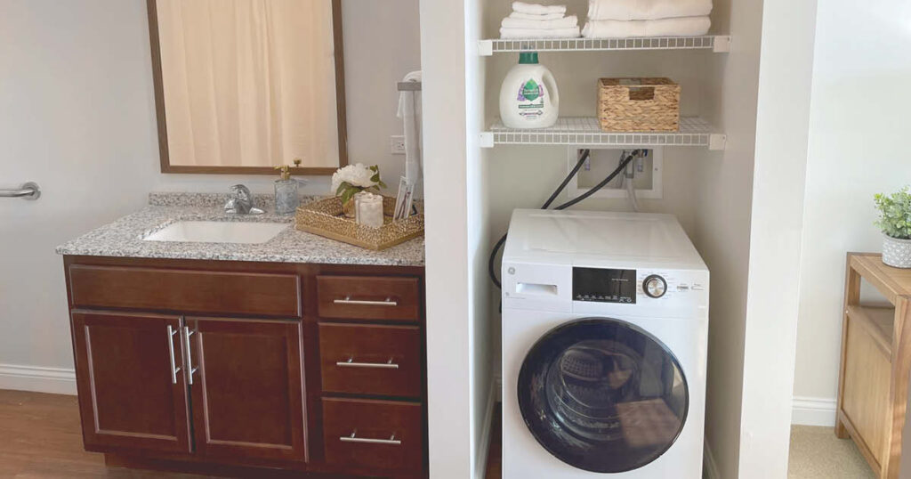 A laundry nook with a front-loading washing machine, detergent, towels, and a wicker basket on white shelves, next to a bathroom vanity with a granite countertop and a mirror.
