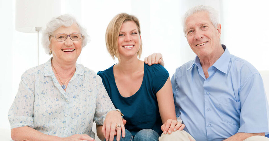 Three people sit close together, smiling at the camera. An elderly woman and man sit on either side of a younger woman, who has her arm around the elderly man. All appear happy and relaxed in a bright, indoor setting.