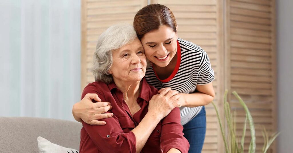 A young woman stands behind an older woman seated on a couch, smiling and hugging her from behind in a cozy, well-lit living room.