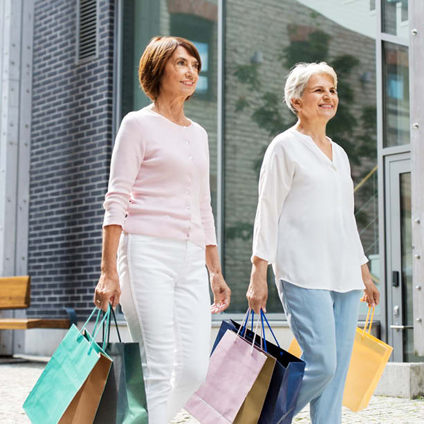 Two smiling older women walk outdoors carrying colorful shopping bags. They are dressed in light, casual clothing, and there is a modern building with large windows and a brick wall in the background.